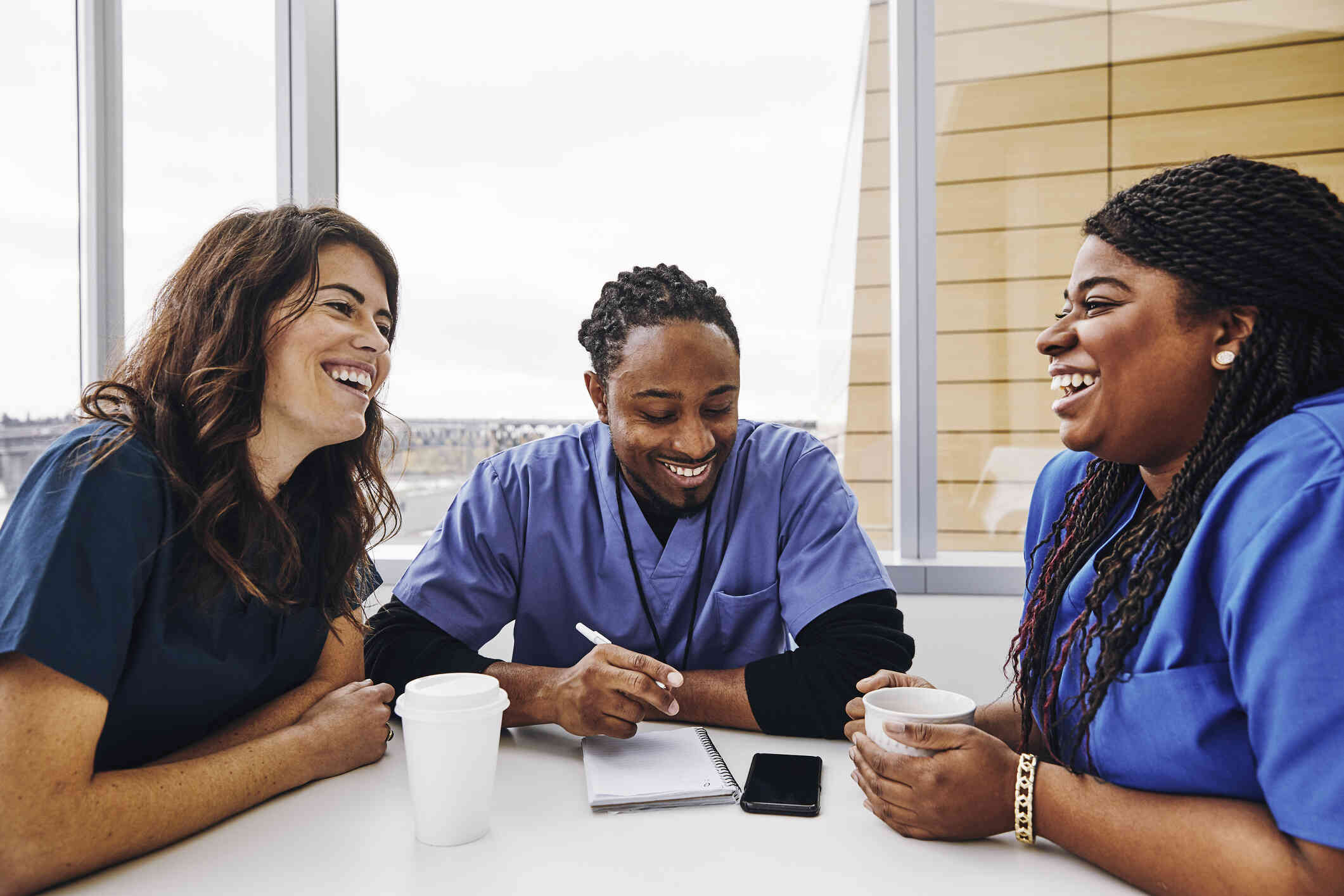 Two women and a man in blue scrubs laugh as they sit at a table together. The man holds a pen and writes in a notebook and the two women have coffee cups in front of them.